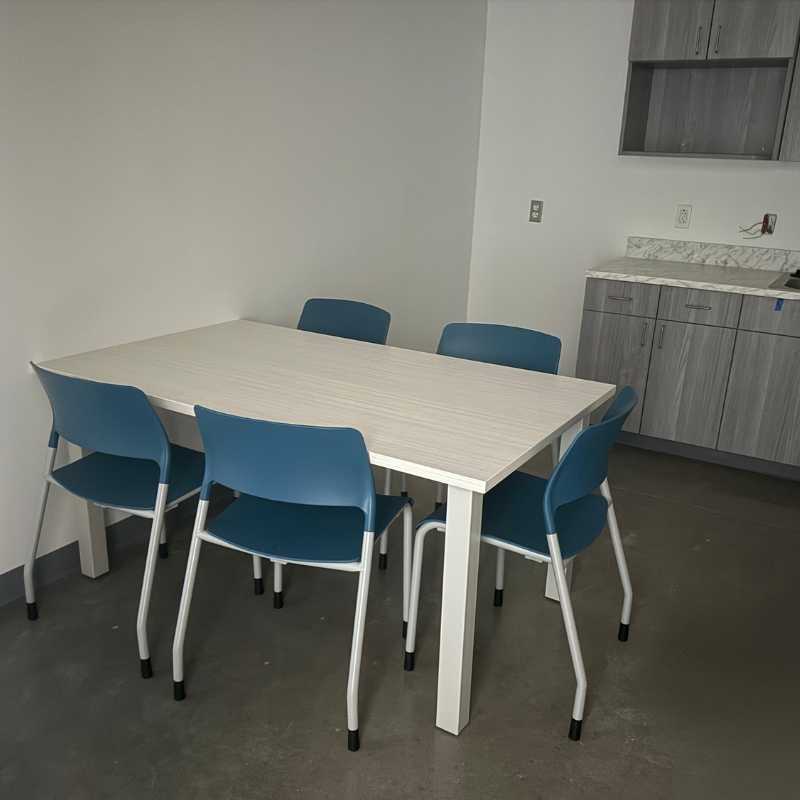 Compact break room with rectangular laminate table, blue stacking chairs, and built-in kitchenette